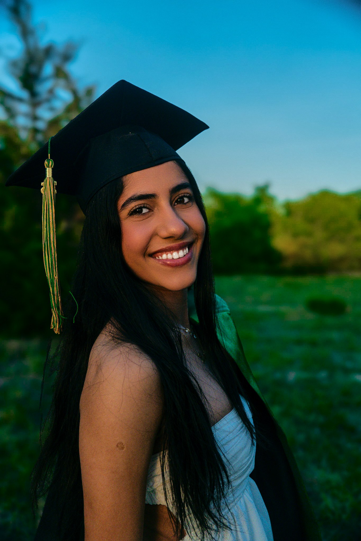 Outdoor graduation portrait in a black cap against green trees.